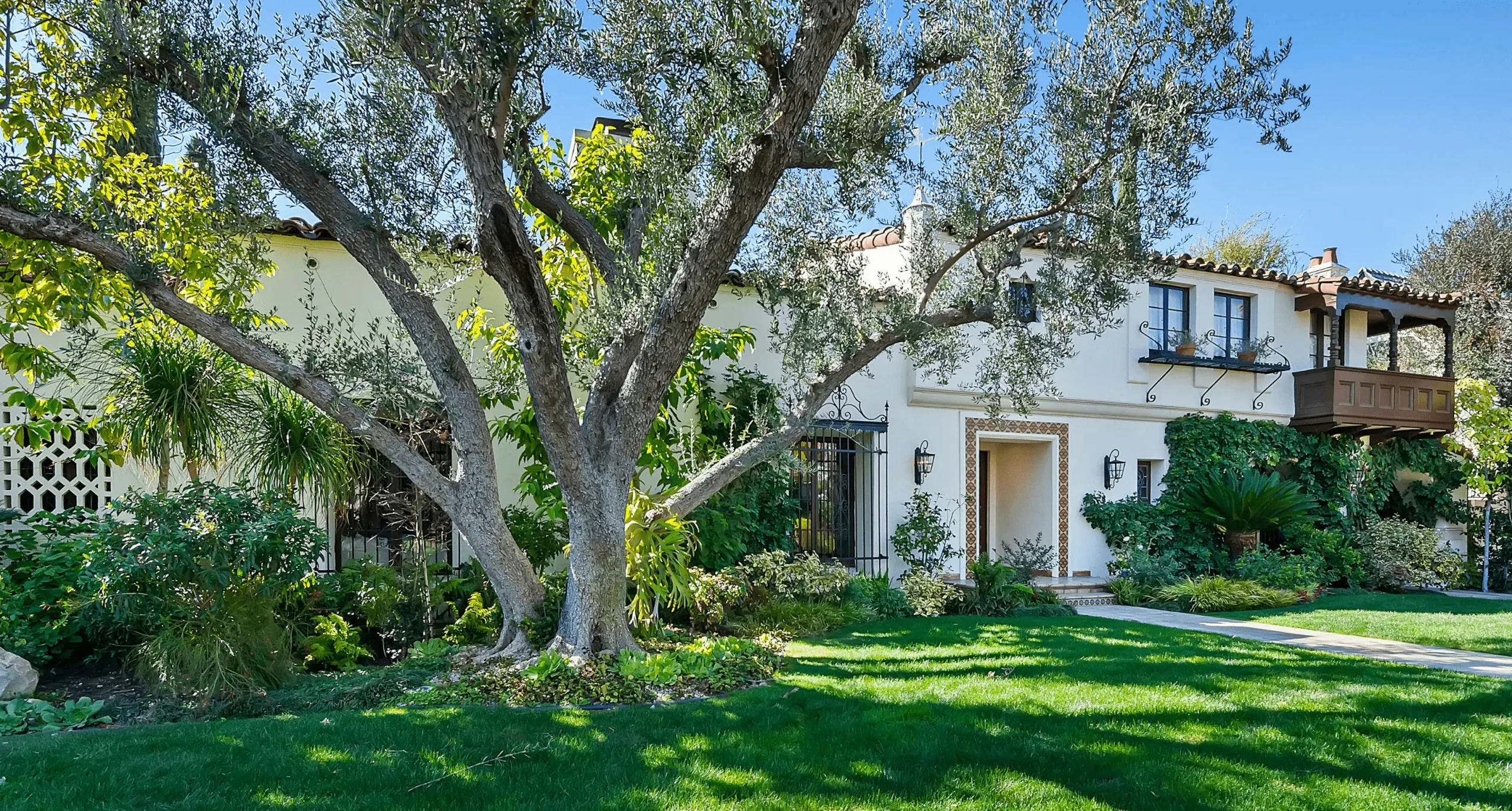 Traditional two-story home with white stucco walls and terracotta roof tiles, featuring ornate black wrought iron balcony railings and a gated entry. Lush landscaping, including mature trees, manicured lawn, and vibrant green shrubs, enhance the property's curb appeal